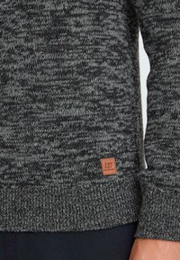Close-up van een grijs en zwart gebreid sweater met ribbels aan de onderrand en mouwen, met een klein bruin lederen label met een logo nabij de onderrand.