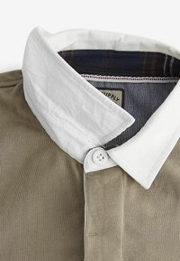 Polo shirt verde oliva con colletto bianco, placca dei bottoni e finitura testurizzata. L'interno presenta un motivo a quadri in colori scuri.