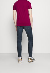 En åtsittande vinröd t-shirt, blå slim-fit jeans och grå-silver sneakers med guldaccenter, sedda bakifrån mot en neutral bakgrund.