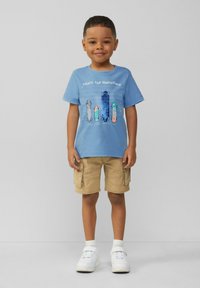 Junger Junge steht, trägt ein T-Shirt mit blauem Skateboard-Graphic, khakifarbene Cargo-Shorts, weiße Socken und weiße Sneaker vor einem schlichten Hintergrund.