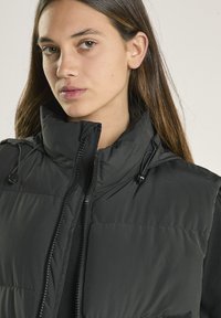 Gilet imbottito nero con collo alto, chiusura lampo e coulisse regolabili. Realizzato in tessuto liscio con imbottitura trapuntata per l'isolamento.