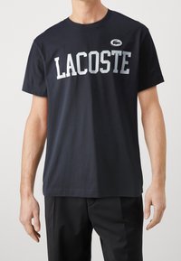 Tummansininen puuvillapaita, jossa on suuri valkoinen "LACOSTE" -painatus ja pieni logo-patch. Klassinen pyöreä kaula-aukko ja lyhyet hihat.