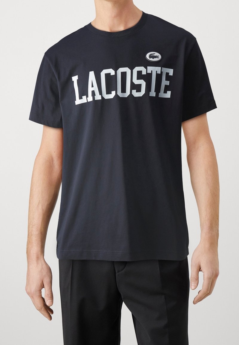 Granatowa bawełniana koszulka z dużym białym napisem "LACOSTE" oraz małą naszywką z logo. Klasyczny krój z okrągłym dekoltem i krótkimi rękawami.