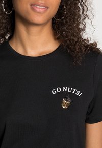 Vrouw in een zwart shirt met gouden en zwarte pailletten eikel en witte tekst "GO NUTS!" dichtbij de linkerschouder.