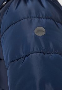 Mørkeblå puffjakkeerm med kviltede seksjoner, med en rund stofflapp merket "DENIM ATTITUDE" i grå. Glatt tekstur.