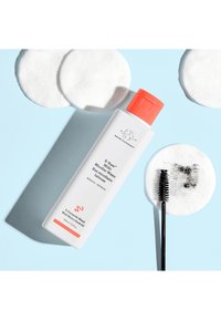 E-Rase Milky Micellar Wasser in einer weißen Flasche mit einem orangefarbenen Deckel, begleitet von Wattepads und einem schwarzen Mascara-Wimpernbürstchen, auf einem blauen Hintergrund.