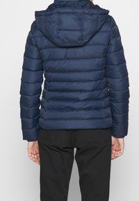 Person som bär en marinblå kviltad pufferjacka med huva, i kombination med svarta byxor, sedd bakifrån mot en enkel bakgrund.