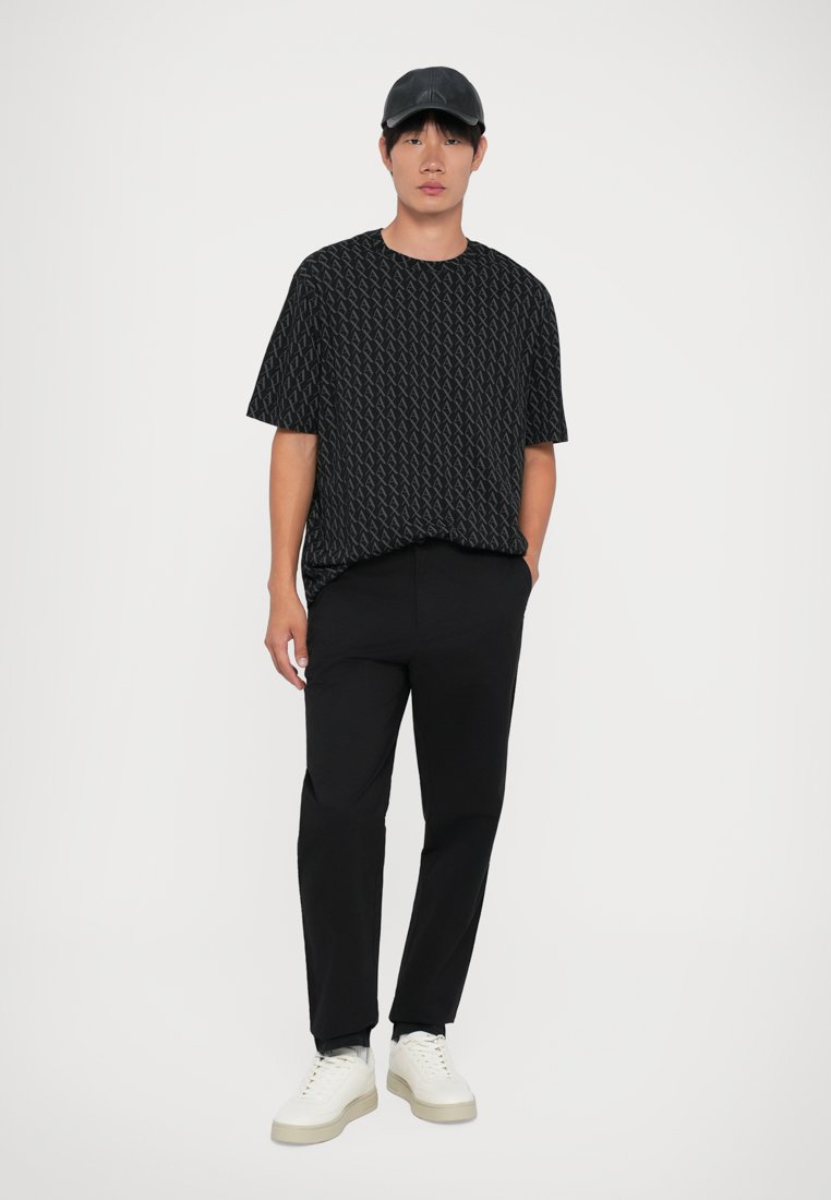 T-shirt noire à manches courtes avec un motif géométrique, associée à un pantalon noir et des baskets blanches. Une casquette noire à visière plate complète le look.