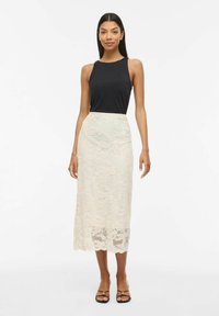 Haut noir sans manches associé à une jupe midi en dentelle ivoire avec des motifs floraux et un ourlet festonné. Le mannequin porte des sandales à enfiler.