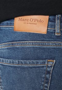 Detalj av en baksida av blå denimjeans med en brun läderetikett från Marc O’Polo sydd på midjan.