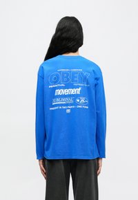 Μπλε μακρυμάνικη μπλούζα με λευκό κείμενο στην πλάτη, που εμφανίζει λογότυπα και φράσεις. Το υλικό φαίνεται μαλακό· εφαρμοστή γραμμή με στρογγυλή λαιμόκοψη.