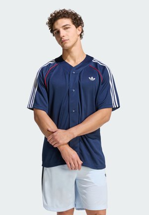 Jaunas vyras su garbanotais plaukais, dėvintis tamsiai mėlyną Adidas sportinę marškinėlę ir baltus šortus, stovintis sukryžiuotomis rankomis priešais šviesią foną.