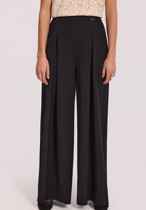 Czarne spodnie o szerokich nogawkach z plisami, gładki materiał i subtelny detal w pasie, zestawione z koronkową bluzką oraz dodatkiem w postaci pierścionka.