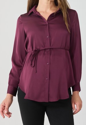 Kvinde iført en langærmet, knappet lilla bluse med tynd taljebånd og sorte bukser, vist fra nakke til hofter.