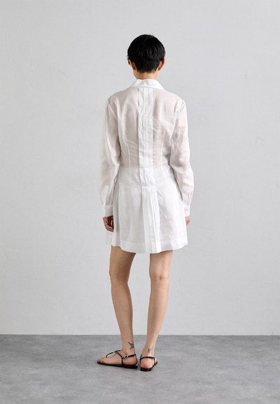 Vestito camicia bianco in lino con maniche lunghe, dettaglio della cucitura sul retro e una vestibilità ampia che arriva sopra le ginocchia, abbinato a sandali neri.