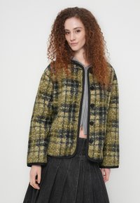 Γυναίκα με σγουρά κόκκινα μαλλιά, φοράει πράσινο και μαύρο καρό μπουφάν με κουμπιά πάνω από γκρι φούτερ με φερμουάρ και σκουρόχρωμη πλισέ φούστα.