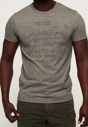 Uomo che indossa una maglietta grigia aderente con trama e testo in rilievo, abbinata a pantaloni cargo verde oliva con tasche e dettagli a toppa.