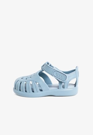 Helle blaue Sandale aus weichem Kunststoff mit einer schnürigen Form und Ausschnitten, verstellbarem Riemen und flacher Sohle.