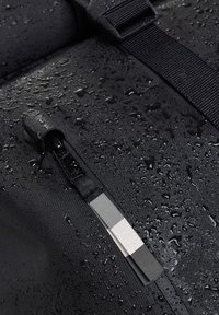Zaino impermeabile nero con superficie strutturata, dotato di una zip e una tracolla resistenti all'acqua. La superficie mostra gocce d'acqua.