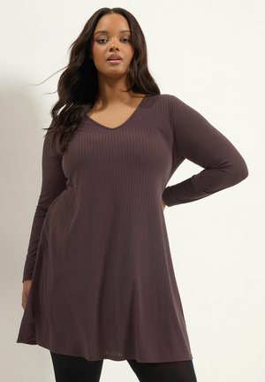 Femme aux longs cheveux foncés portant une robe côtelée à manches longues et col en V, de couleur violet-brun, et des leggings noirs, debout avec une main sur la hanche.
