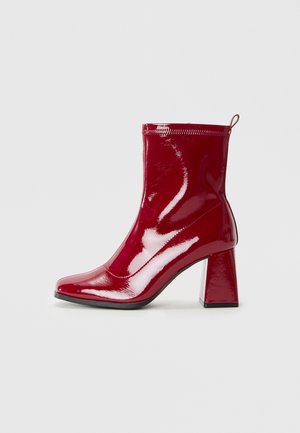 Botines de charol rojo con punta cuadrada y tacón grueso. Cuentan con un borde superior con cremallera y una tirador en la parte trasera para facilitar su uso.