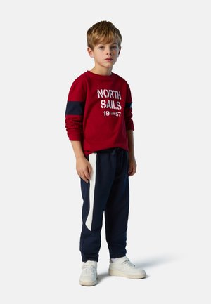 Roter Pullover mit "NORTH SAILS 1957" in weißer Schrift, kombiniert mit marineblauen Jogginghosen mit einem weißen Seitestreifen. Weiße Sneakers runden den Look ab.