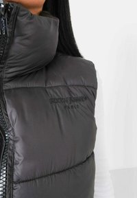 Schwarze Steppweste mit hohem Kragen; verfügt über ein gestepptes Design, geprägtes Logo in Schwarz und einen Reißverschluss vorne. Glatte, glänzende Textur.