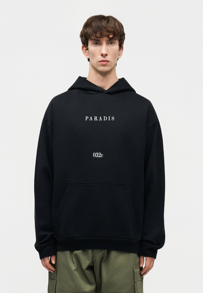 Jeune homme portant un sweat à capuche noir avec les inscriptions "PARADIS" et "032c" sur le devant, associé à un pantalon cargo vert olive, debout devant un fond blanc.