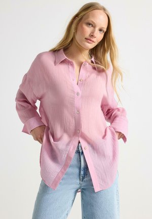 Kvinde iført løs, lyserød skjorte med knapper og lyseblå jeans, poserer med hænderne i lommerne mod en ensfarvet hvid baggrund.