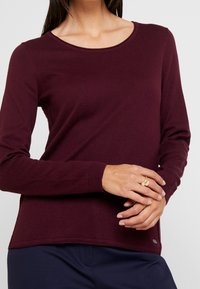 Femme portant un pull bordeaux à manches longues et un pantalon sombre, les mains jointes et une bague en or au doigt.