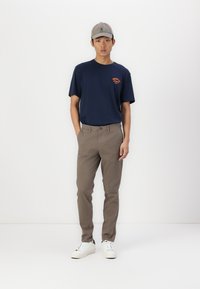Marineblaues T-Shirt mit orangefarbenem Logo auf der Brust, beigen Chinos und weißen Sneakers. Das Modell trägt eine graue Mütze und steht vor einem einfarbigen Hintergrund.