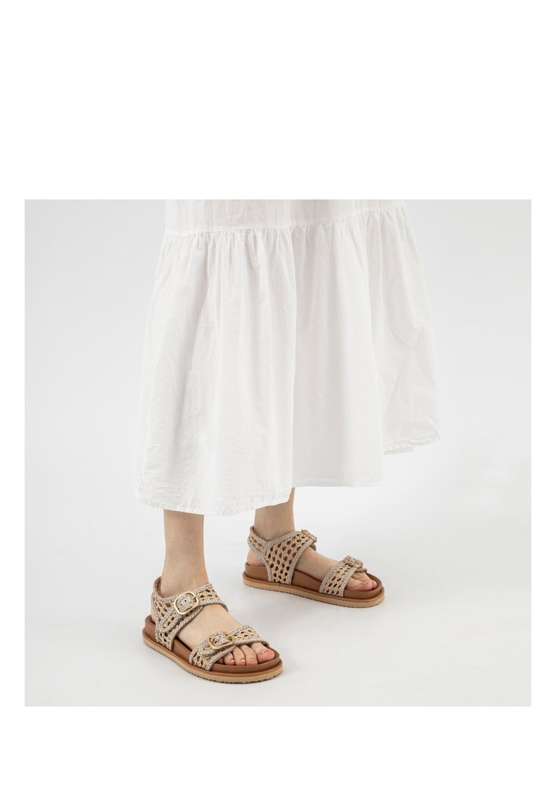 Feet wearing beige woven sandals with buckles, paired with a white flowing skirt against a plain light background.