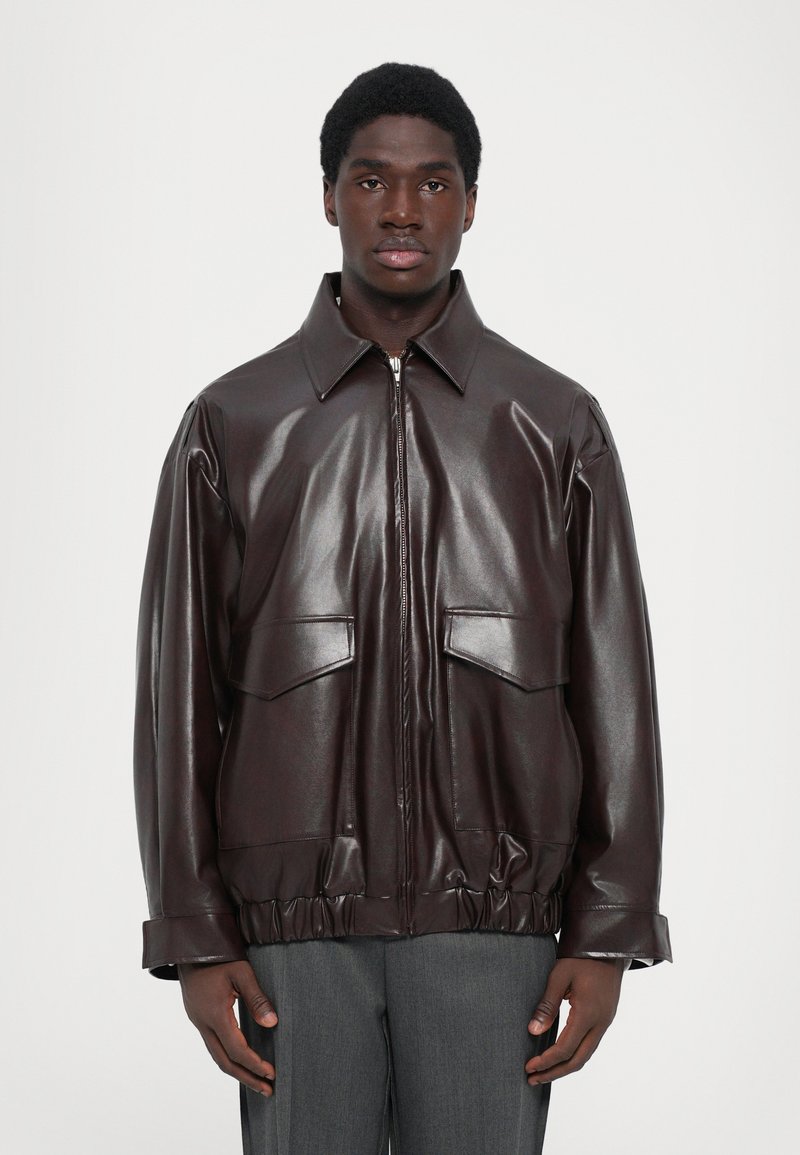 Giacca in pelle marrone con zip frontale, grandi tasche sul petto e orlo arricciato. Presenta un colletto classico e una texture liscia e lucida.