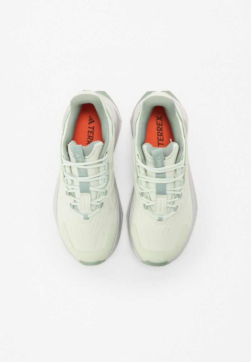 Zapatillas deportivas de color verde claro con parte superior de textil, detalles de malla y una suela acolchada. Presenta un cierre de cordones y una plantilla con el logo en naranja.
