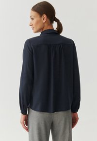Marineblauwe blouse met lange mouwen, een platte kraag en een rugplooinstructie, gemaakt van een gladde stof. Mouwen met manchetten en een rechte zoom.