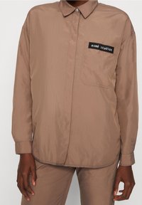 Hnědá košile s dlouhým rukávem a přední kapsou, na které je černá nálepka "esmé studios", nošená s odpovídajícími hnědými kalhotami na osobě s tmavou pleti.