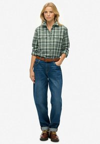 Blonde Frau trägt ein grünes karriertes Hemd, blaue, hochgekrempelte Jeans mit braunem Gürtel und braunen Schnürboots und steht mit einer Hand in der Tasche.