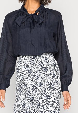 Vrouw die een zwarte getextureerde blouse draagt met een strik bij de hals en een witte rok met zwarte dierenprint.