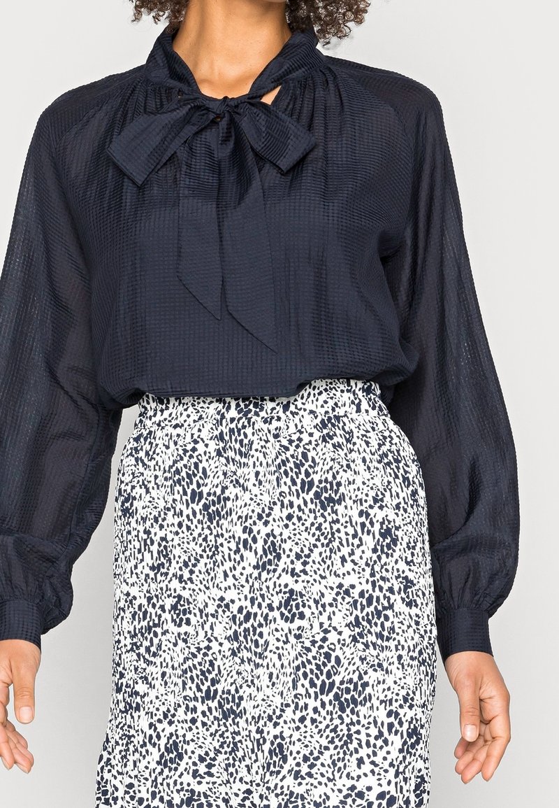 Femme portant un chemisier noir texturé avec un nœud au cou et une jupe blanche avec un imprimé animal noir.