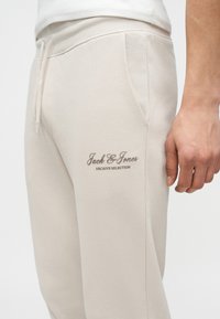 Μπεζ καθημερινό παντελόνι με πλαϊνή τσέπη και κορδόνι στη μέση, με το λογότυπο "Jack & Jones Archive Selection" κοντά στον επάνω μηρό.