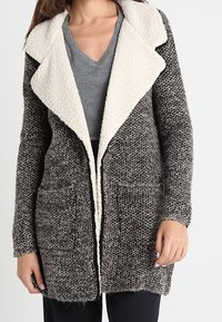 Stickad cardigan med ett strukturerat grått och svart mönster, kontrasterande krämfärgad fuskpäls-krage och framfickor. Öppen framdesign.