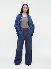 Femme aux longs cheveux foncés portant un jean bleu foncé ample, un débardeur gris, une veste en jean bleue déboutonnée et des chaussures noires à bout pointu.