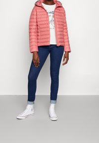 Mujer vistiendo una chaqueta acolchada de color coral, camiseta gráfica blanca, jeans ajustados oscuros remangados y zapatillas altas blancas, de pie contra una pared blanca.