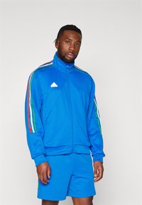 Tuta sportiva blu in materiale liscio, caratterizzata da una giacca con zip completa e righe multicolori, abbinata a pantaloni corti.