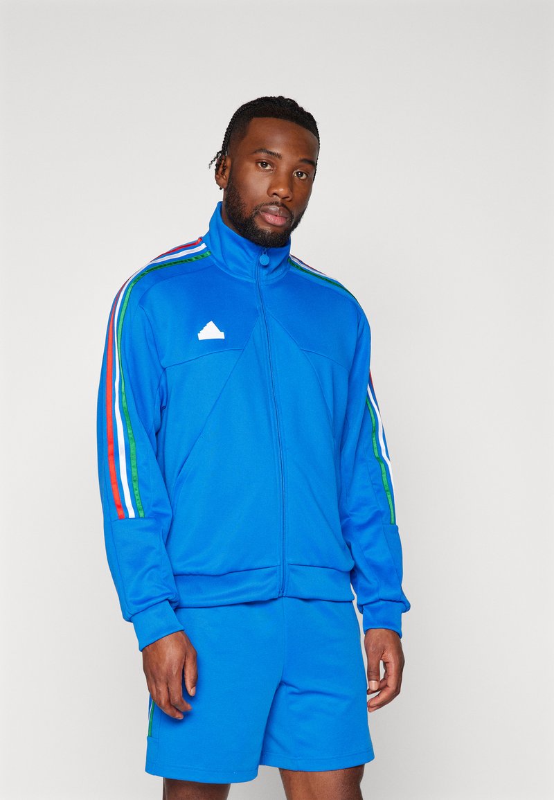 Tuta sportiva blu in materiale liscio, caratterizzata da una giacca con zip completa e righe multicolori, abbinata a pantaloni corti.