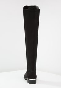 Bota plana negra por encima de la rodilla con parte superior lisa, punta redonda, costura trasera y tacón bajo apilado con acento plateado sobre una superficie blanca.