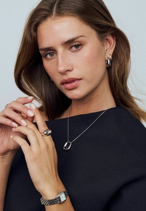 Smycken i silver inkluderar ett hängande halsband och en stor ring, samt en klocka med texturerad metallyta. Modellen har långt hår och mjuk makeup mot en mörk topp.