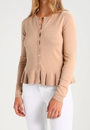 Vrouw draagt een beige, knoopjesvest met een gerimpelde zoom en lange mouwen, gecombineerd met een witte broek, staand tegen een effen achtergrond.