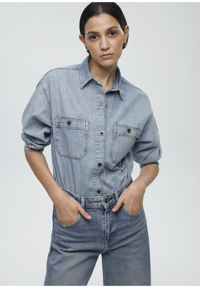 Denim shirt in light blue, featuring two chest pockets with button closures, relaxed fit, and contrasting stitching. Tucked into high-waisted jeans.