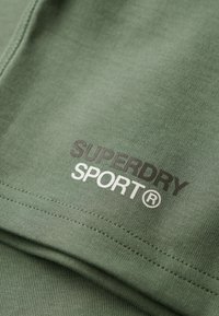 Tessuto di cotone verde con i loghi "SUPERDRY" e "SPORT" in bianco e nero. La texture è liscia con dettagli di cucitura visibili.